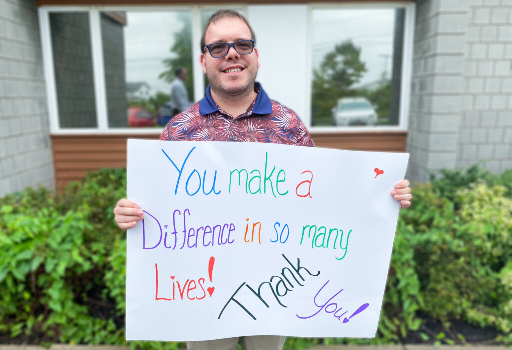 A man holds a sign reading, “You make a difference in many lives,” smiling warmly at the camera. A man holds a sign reading, "You make a difference in many lives," smiling warmly at the camera.
